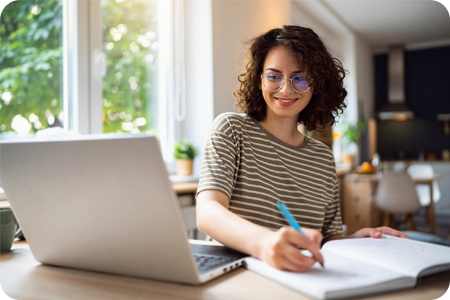 Young female learner writing in a note pad near a laptop
